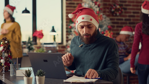 Male employee wearing santa hat in festive office  working on laptop to send email before christmas party celebration. Businessman using pc at desk during winter holiday season  xmas decor.
