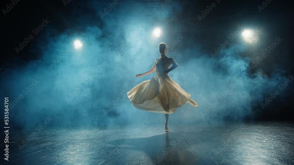Graceful ballerina dances in light flowing dress on stage in pointlight in stage smoke. Ballet dancer performs dance elements of classical ballet