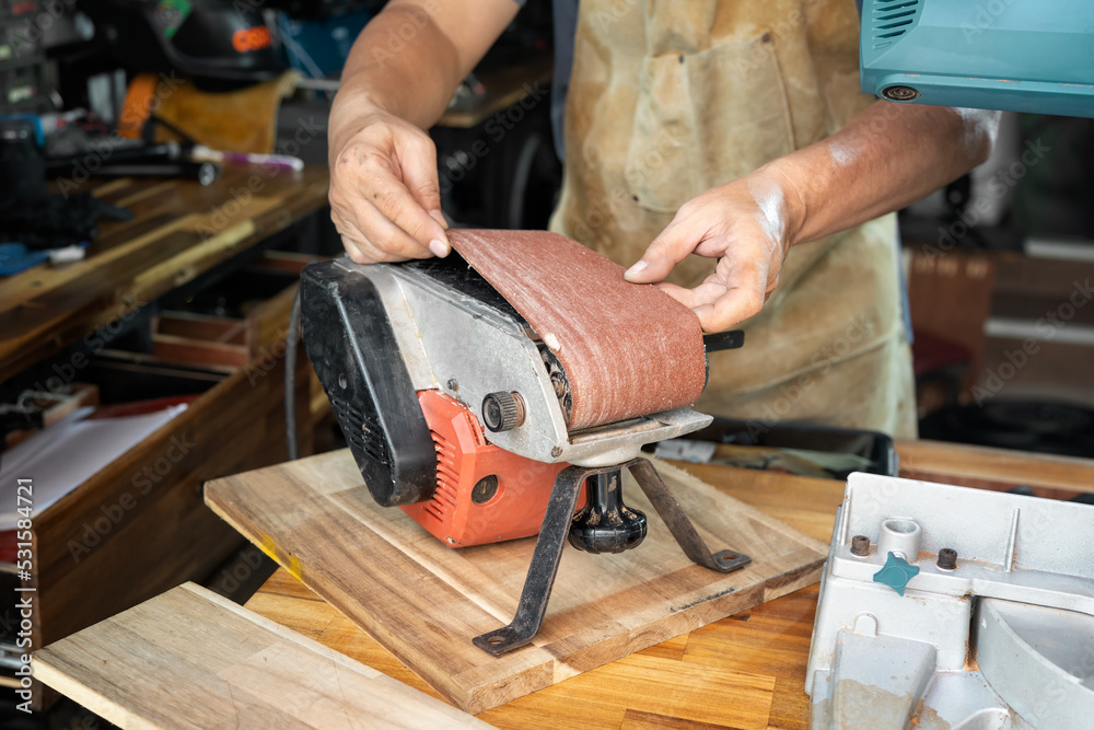 carpenter wearing apron and Install or remove abrasive belt of Belt Sander on wooden tabel in the workshop ,DIY maker and woodworking concept. selective focus carpenter wearing apron and Install or remove abrasive belt of Belt Sander on wooden tabel in the workshop ,DIY maker and woodworking concept. selective focus
