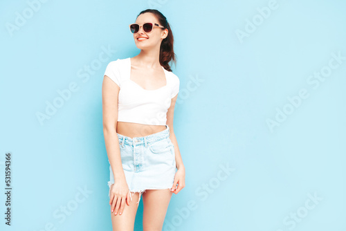 Young beautiful smiling female in trendy summer jeans skirt and top  clothes. Sexy carefree woman posing near blue wall in studio. Positive brunette model having fun.Cheerful and happy. In sunglasses © halayalex