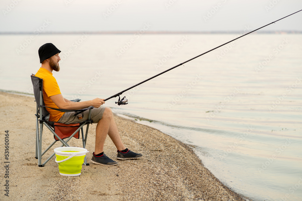 Millennial man fishing sitting on chair on lake in Finland at summer. Recreational fishing fisherman outdoor leisure. Millennial man fishing sitting on chair on lake in Finland at summer. Recreational fishing fisherman outdoor leisure.