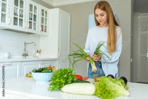 Young woman split green onion above table full of greens and vegetables. Housewife make salad for dinner. Organic products without nitrates. Vegetarianism, proper nutrition, healthy eating, cooking.