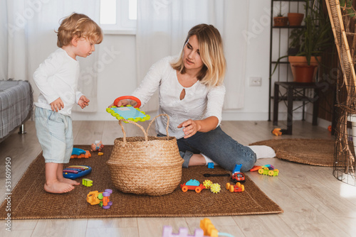 Young woman, housewife puts toys in the basket with her little son at home