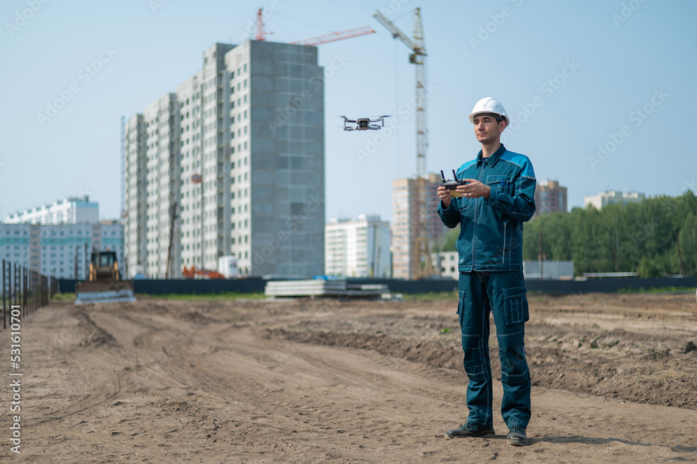 A man in a helmet and overalls controls a drone at a construction site. The builder carries out technical oversight.