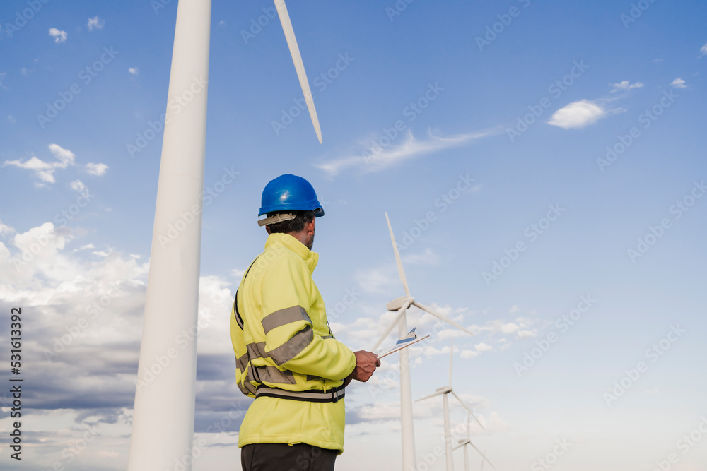Engineer wearing reflective clothing looking at wind turbines by sky Engineer wearing reflective clothing looking at wind turbines by sky