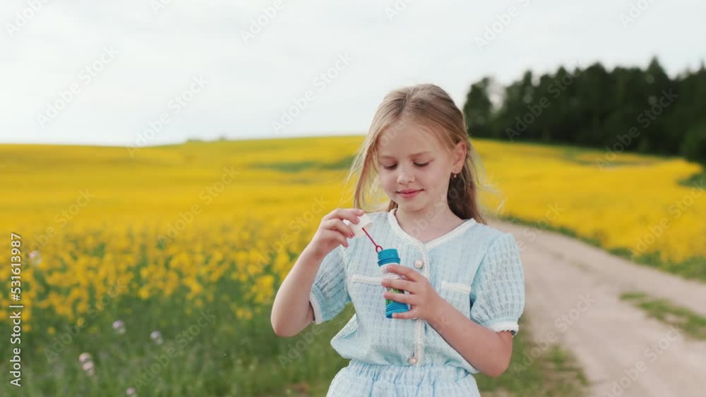 Cheerful caucasian girl in beautiful summer dress blowing soap bubbles while standing among countryside with yellow field on background. Free time with fun outdoors. Cheerful caucasian girl in beautiful summer dress blowing soap bubbles while standing among countryside with yellow field on background. Free time with fun outdoors.
