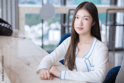 Asian young woman with long hair while she sits on chair in workplace with glassed building as a background.