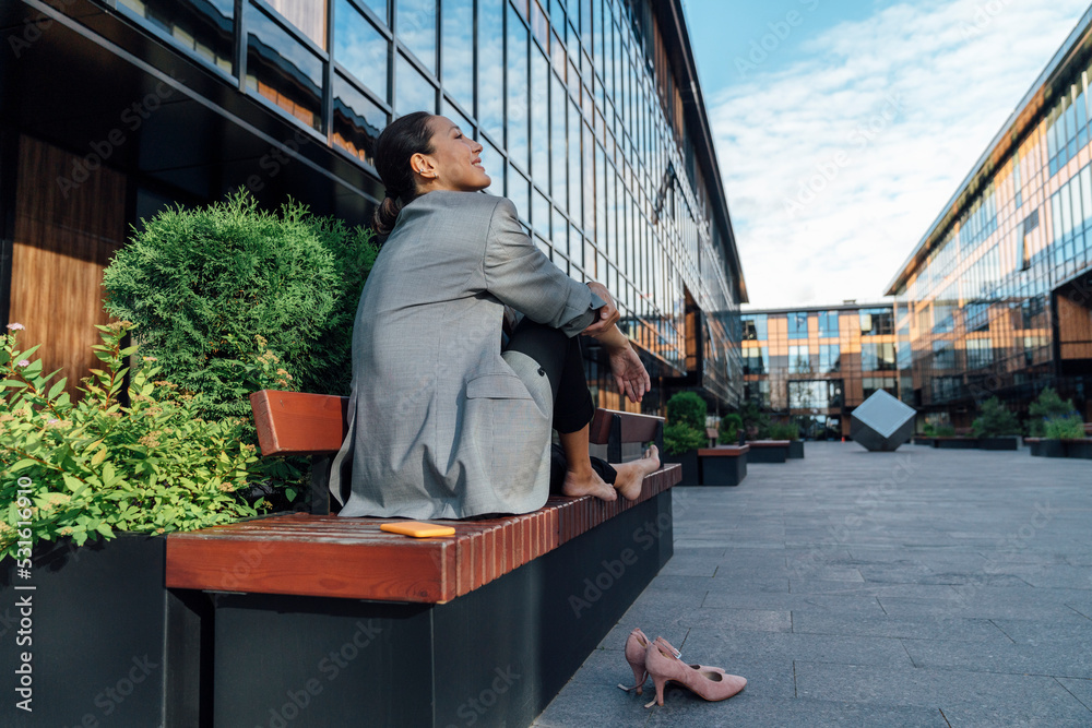 Smiling businesswoman sitting on bench in front of office building Smiling businesswoman sitting on bench in front of office building
