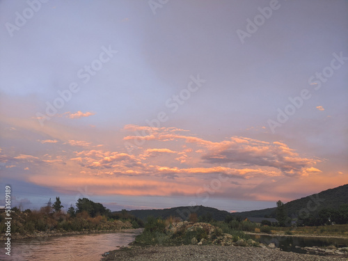 Beautiful cloudy sky at sunset over the river. The high bank of the river is overgrown with grass.