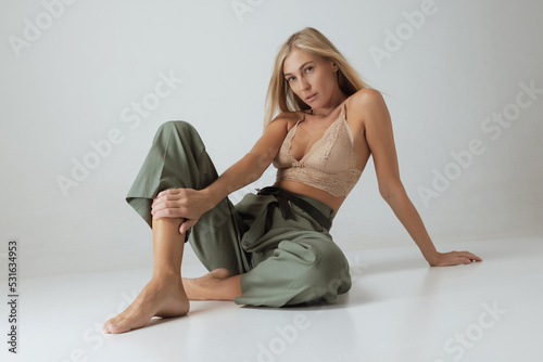 Beautiful woman with long blond hair and natural makeup wearing lacy top and casual trousers sitting on floor isolated on grey background.