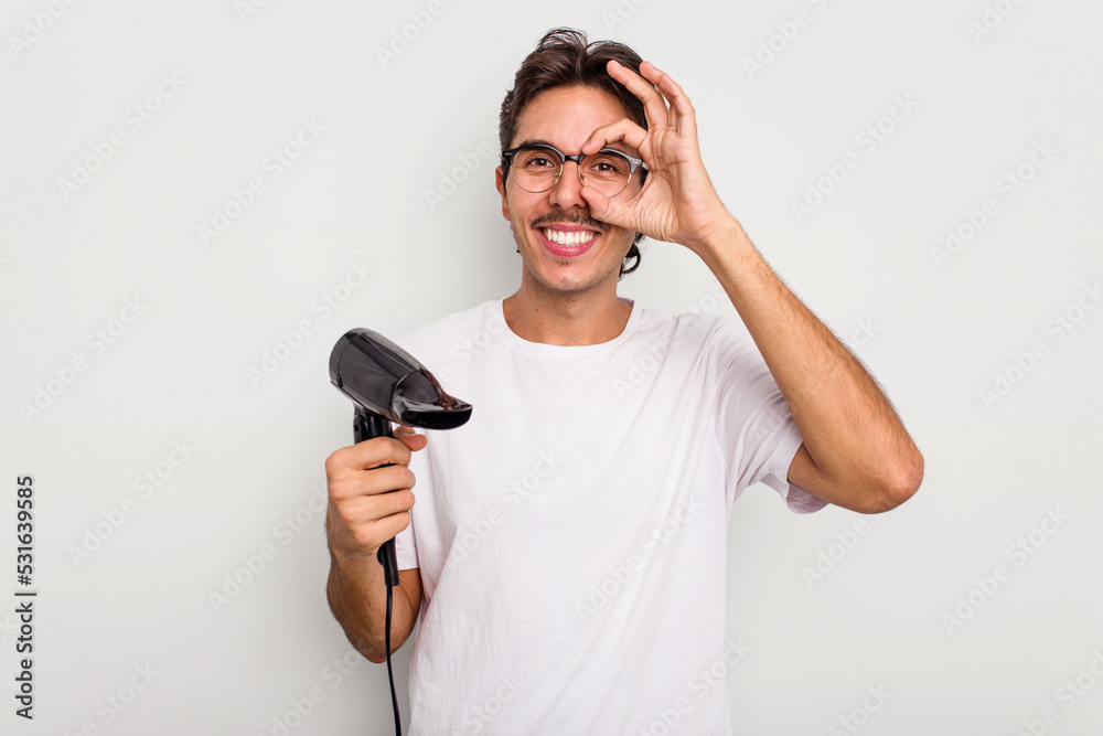 Young hispanic man holding a hairdryer isolated on white background excited keeping ok gesture on eye. Young hispanic man holding a hairdryer isolated on white background excited keeping ok gesture on eye.