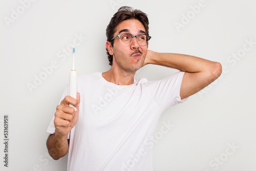 Young hispanic man holding electric toothbrush isolated on white background touching back of head, thinking and making a choice.