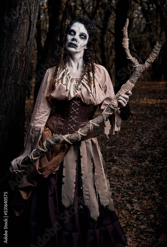 Halloween (Samhain) theme: ugly terrible voodoo witch with staff. Portrait of evil hag in dark forest. Zombie woman (undead)
