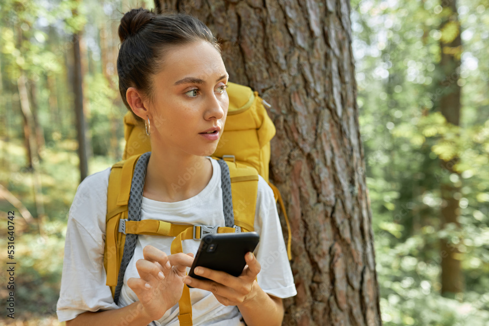 Girl tourist with combed hair and yellow backpack on shoulders lost in forest, holding smartphone, dialing number of her friend, using gps navigation system, trying to catch signal of mobile network