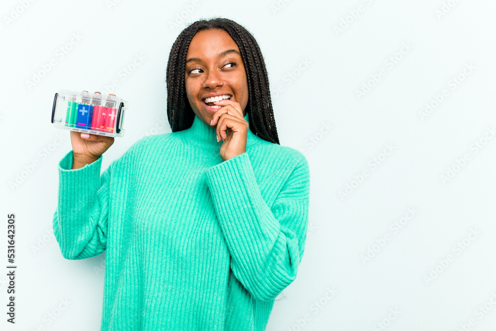 Young African American woman holding battery box isolated on blue background relaxed thinking about something looking at a copy space. Young African American woman holding battery box isolated on blue background relaxed thinking about something looking at a copy space.