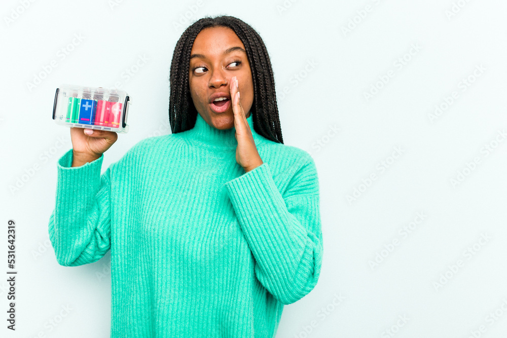 Young African American woman holding battery box isolated on blue background is saying a secret hot braking news and looking aside Young African American woman holding battery box isolated on blue background is saying a secret hot braking news and looking aside