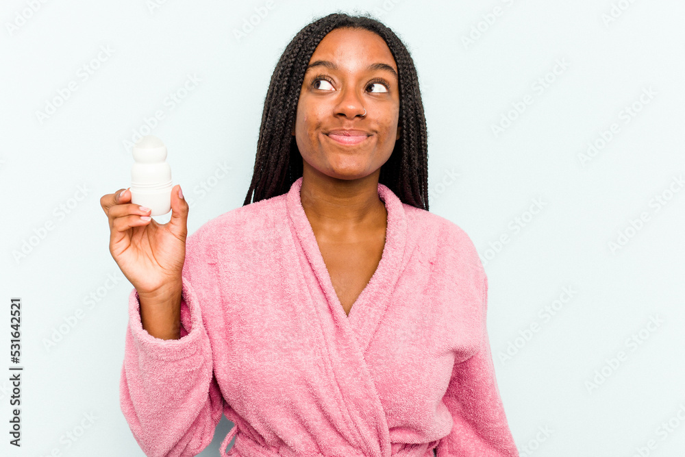 Young African American woman wearing bathrobe holding a deodorant isolated on blue background dreaming of achieving goals and purposes Young African American woman wearing bathrobe holding a deodorant isolated on blue background dreaming of achieving goals and purposes