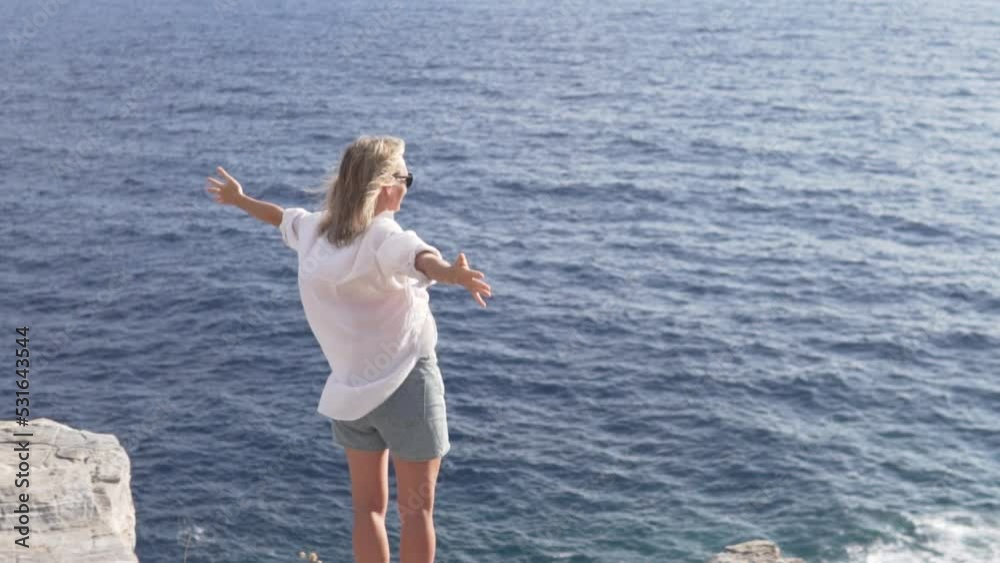 View from the back. A female tourist raises her hands up on a rock by the sea. white shirt develops in the wind View from the back. A female tourist raises her hands up on a rock by the sea. white shirt develops in the wind