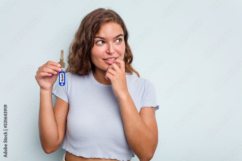 Young caucasian woman holding home keys isolated on blue background relaxed thinking about something looking at a copy space. Young caucasian woman holding home keys isolated on blue background relaxed thinking about something looking at a copy space.