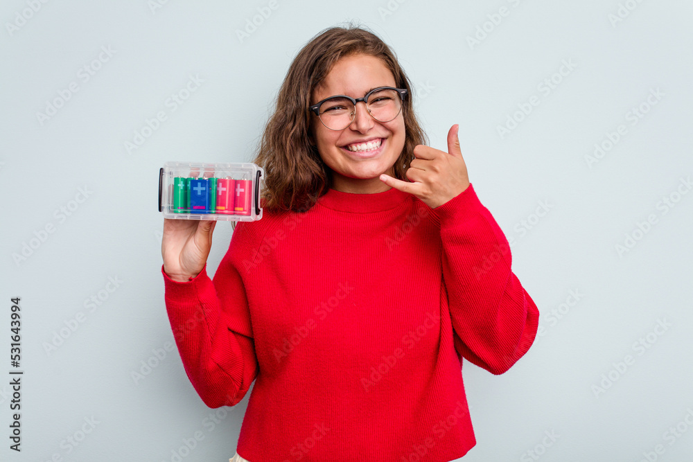 Young caucasian woman holding battery box isolated on blue background showing a mobile phone call gesture with fingers. Young caucasian woman holding battery box isolated on blue background showing a mobile phone call gesture with fingers.