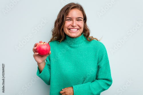 Young caucasian woman with an apple isolated on blue background laughing and having fun.