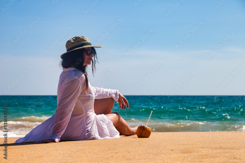 Rear view adult woman in white beachwear, hat and sunglasses with coconut, looking away, sitting on tropical sandy beach at waves ocean background. Travel tourism vacation concept. Copy text space