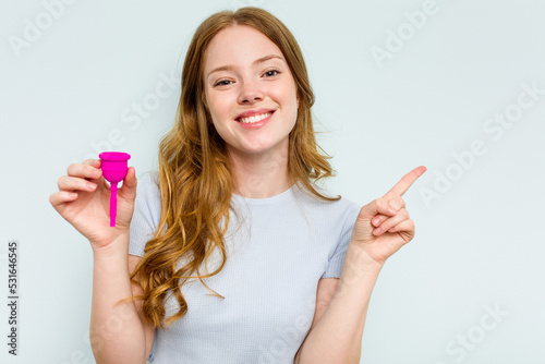 Young caucasian woman holding menstrual cup isolated on blue background smiling and pointing aside, showing something at blank space.