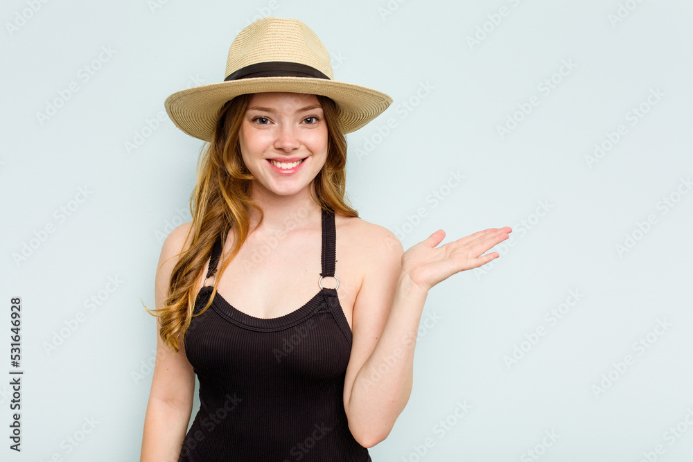 Young caucasian woman going to the beach isolated on blue background showing a copy space on a palm and holding another hand on waist. Young caucasian woman going to the beach isolated on blue background showing a copy space on a palm and holding another hand on waist.