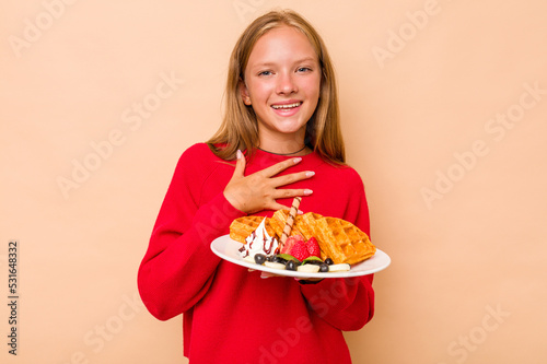 Little caucasian girl holding a waffles isolated on beige background laughs out loudly keeping hand on chest.