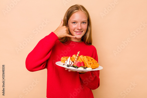 Little caucasian girl holding a waffles isolated on beige background showing a mobile phone call gesture with fingers.