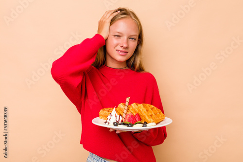 Little caucasian girl holding a waffles isolated on beige background being shocked, she has remembered important meeting.