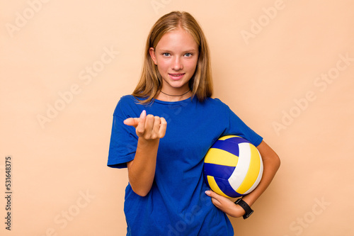 Little caucasian girl playing volleyball isolated on beige background pointing with finger at you as if inviting come closer.