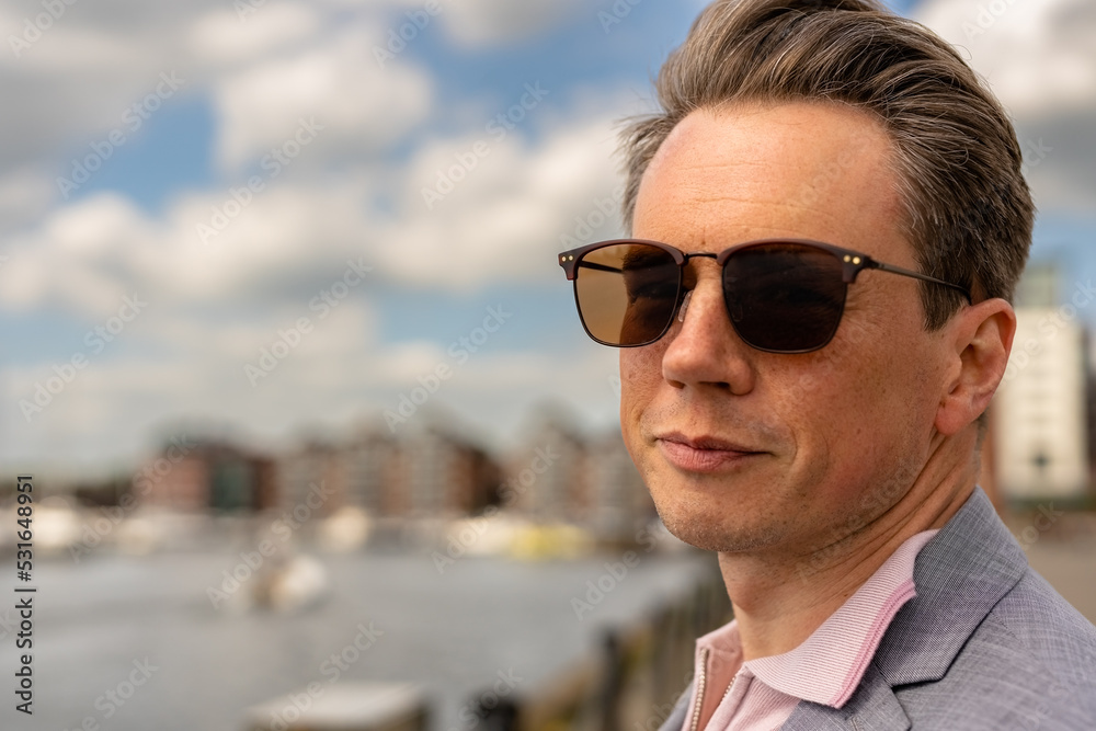 Close up portrait of a handsome businessman in a casual grey suit in front of a busy marina with luxury yachts moored up