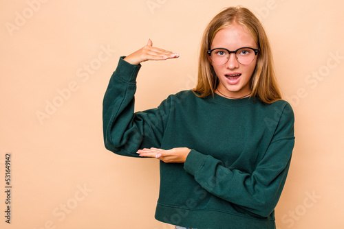 Caucasian teen girl isolated on beige background shocked and amazed holding a copy space between hands.