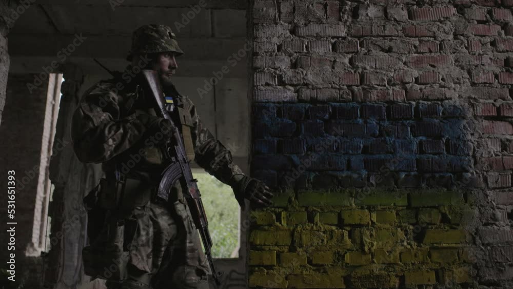 A Ukrainian military man with a weapon in his hands stands against the background of the flag of Ukraine. The flag of Ukraine is painted on a brick wall. A Ukrainian military man with a weapon in his hands stands against the background of the flag of Ukraine. The flag of Ukraine is painted on a brick wall.