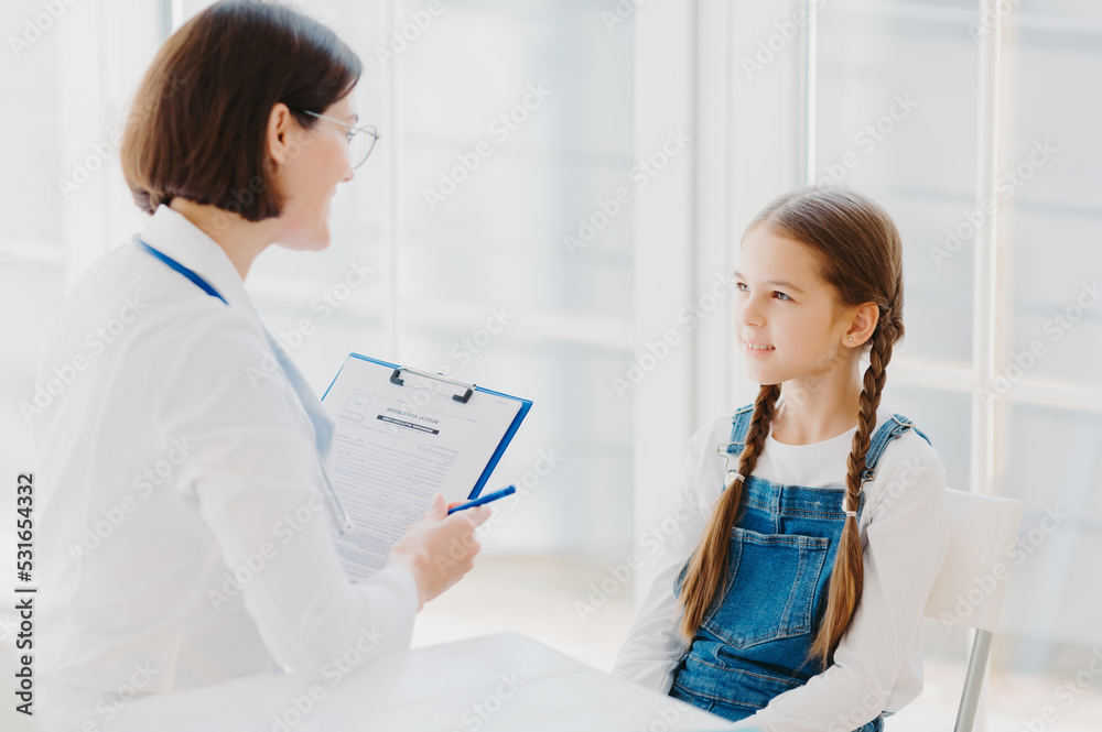 Back view of attentive female pediatrician listens carefully child complaints, writes down notes in personal patient form, gives advice how to cure cold, sit opposite each other in modern clinic