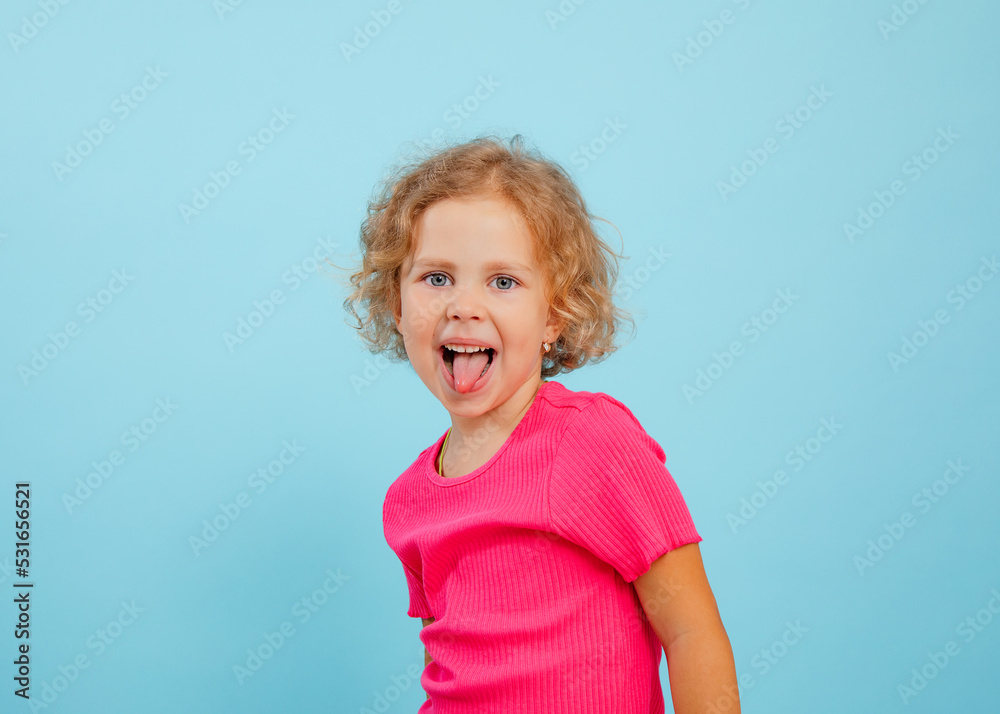 Portrait of happy blue-eyed little girl with curly hair looking, showing tongue, grimacing teasing on blue background. Portrait of happy blue-eyed little girl with curly hair looking, showing tongue, grimacing teasing on blue background.