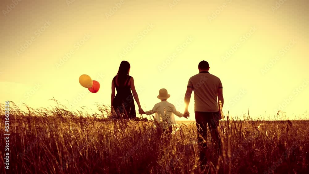 Family walks on field in summer and holds hands, dad speaks and with gesture of hand into distance. Mom carries colored balloons in hands. Happy childhood boy with loved parents. Weekend outdoor. Family walks on field in summer and holds hands, dad speaks and with gesture of hand into distance. Mom carries colored balloons in hands. Happy childhood boy with loved parents. Weekend outdoor.