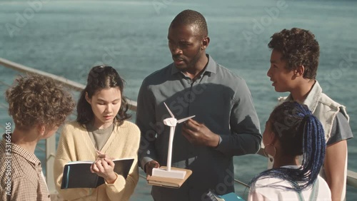 Medium slowmo of group of diverse elementary schoolchildren studying alternative energies outdoors listening to their modern African American male teacher holding windmill model photo