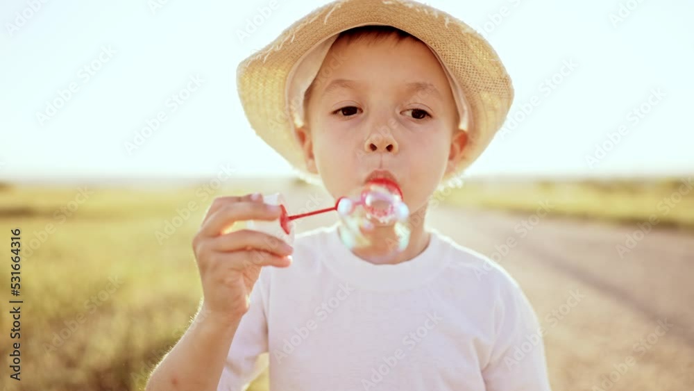 Close up portrait baby boy stands in straw hat on country road in field in summer under the sun rays and blows soap bubbles with rainbow reflection. Games in childhood outdoor on leisure activity. Close up portrait baby boy stands in straw hat on country road in field in summer under the sun rays and blows soap bubbles with rainbow reflection. Games in childhood outdoor on leisure activity.
