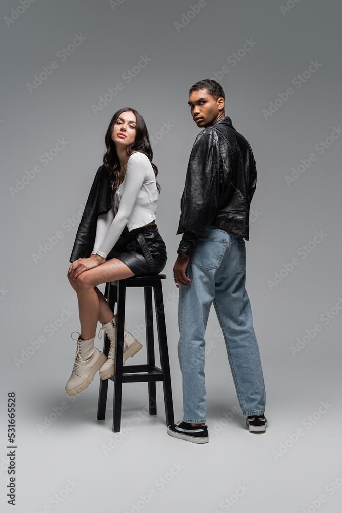 Obraz premium full length of woman in trendy outfit sitting on high chair near stylish african american man on grey.