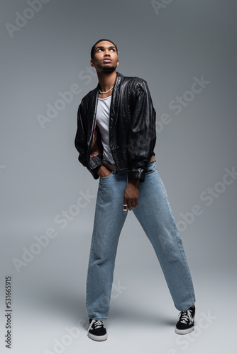 full length of stylish african american man in autumnal outfit posing with hand in pocket of jeans on grey.