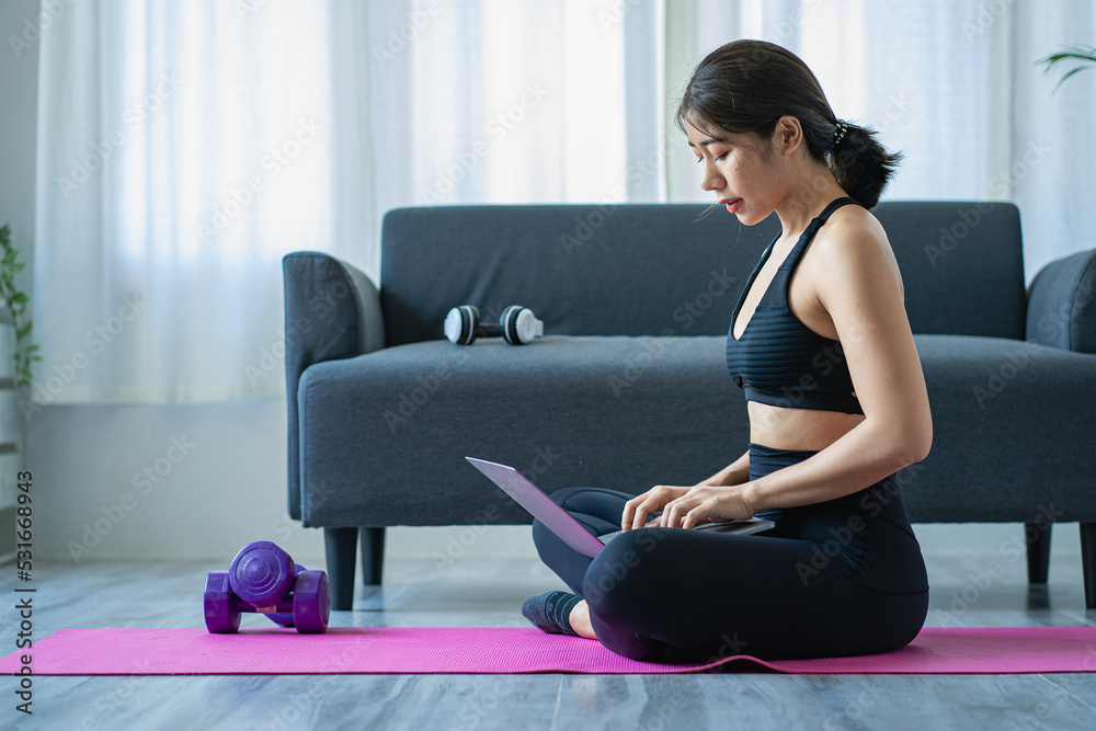 Asian woman in sportswear sitting relaxing and practicing yoga and exercising with dumbbells and laptop computer in bedroom at home fitness concept