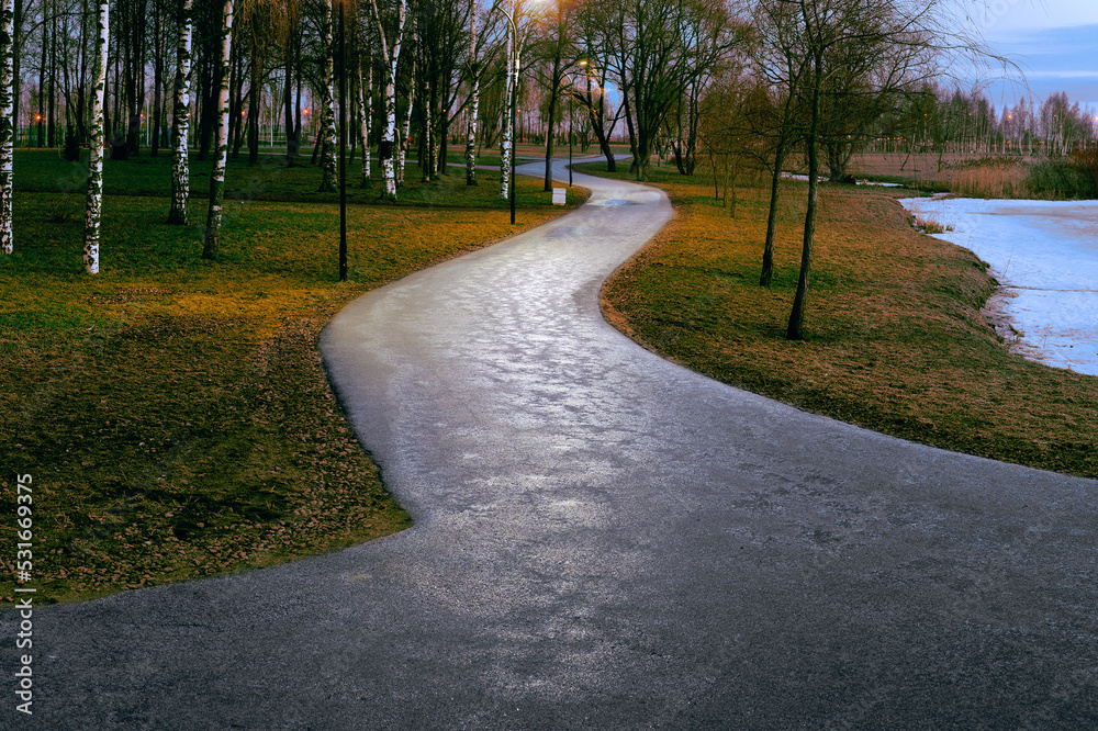 Asphalt gray road in the park at dusk, illuminated by street lamps, meanders between the trees like a huge snake
