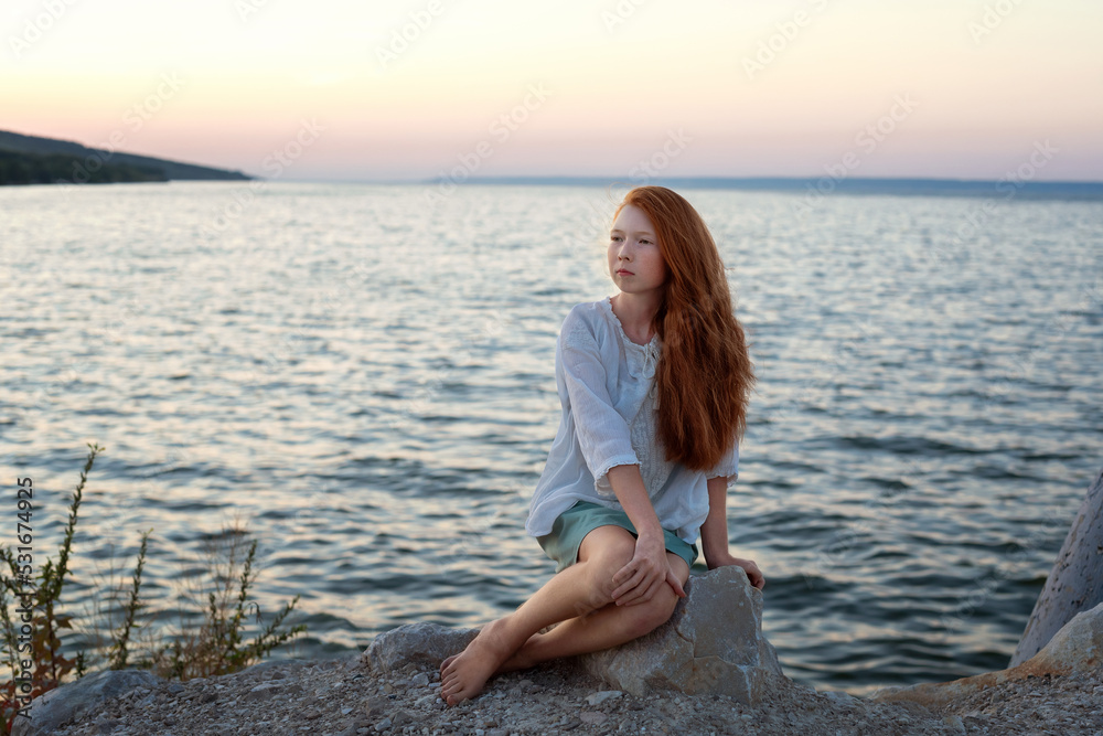A beautiful red-haired girl sits on a stone on the banks of a river, lake, sea. She is looking at ocean and thinking dreamily. Girl alone outside. Girl sitting on rocks. Lonely person.