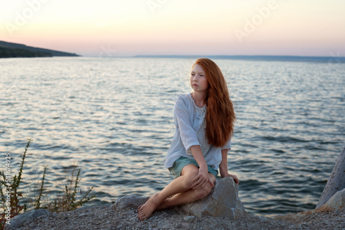 A beautiful red-haired girl sits on a stone on the banks of a river, lake, sea. She is looking at ocean and thinking dreamily. Girl alone outside. Girl sitting on rocks. Lonely person. © Анна Перфилова