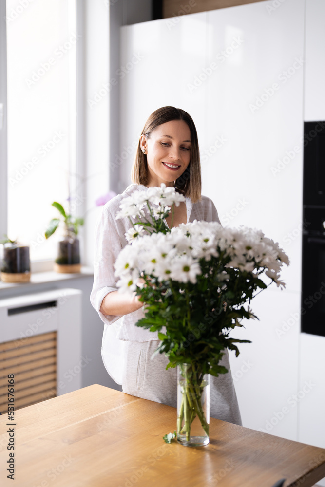 Happy and joyful young woman in white arranging white flowers at home in the kitchen Happy and joyful young woman in white arranging white flowers at home in the kitchen