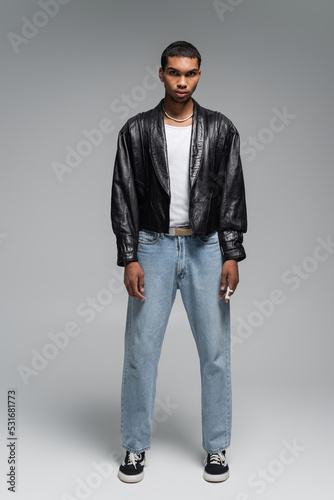 full length of young african american man in jeans and leather jacket on grey.