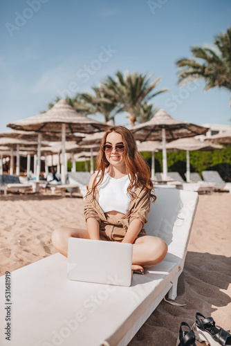 A cheerful blogger girl remotely works with a laptop on the beach with palm trees. A woman makes a video from a trip. Travels the world and works with social networks online. Remote modern profession