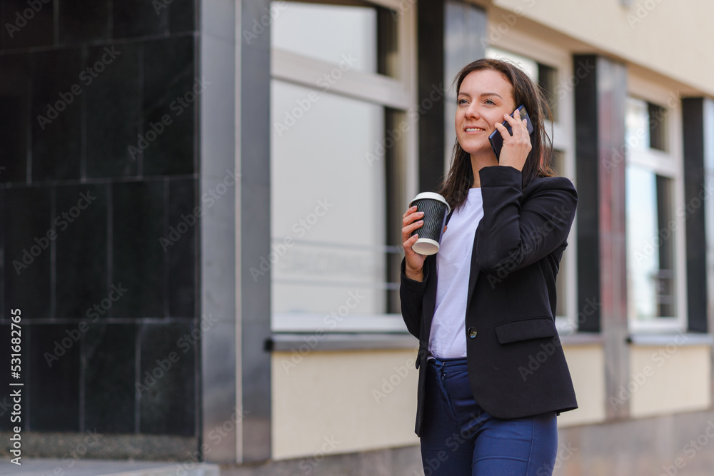 Business woman with coffe paper cup talking on the phone near office.Businesswoman wears a black suit and a white T-shirt.Office building blurred background.Coffe time concept.Summer day. Business woman with coffe paper cup talking on the phone near office.Businesswoman wears a black suit and a white T-shirt.Office building blurred background.Coffe time concept.Summer day.